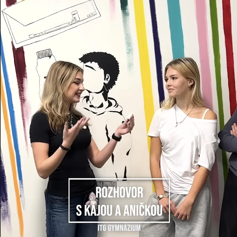 Rozhovor dvou studentek před barevnou nástěnnou malbou.